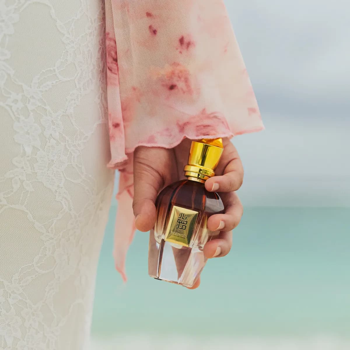 A clear Barfume bottle held against a pink shawl with the lagoon behind